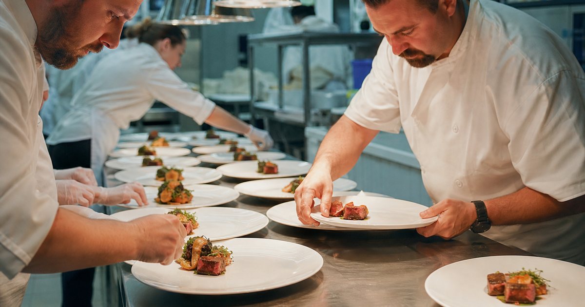 Catering staff in white jackets plating rows of dishes on a stainless steel pass in a busy banquet kitchen, a chef inspecting a plate, heat lamps glowing