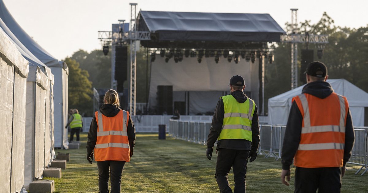 Outdoor festival grounds in early morning light with staff in work vests walking between white tents toward a main stage, dew on the grass, empty barriers